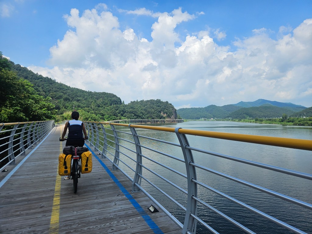 Cycliste sur une véloroute en Corée du Sud, traversant un pont avec des montagnes en arrière-plan et un ciel nuageux.