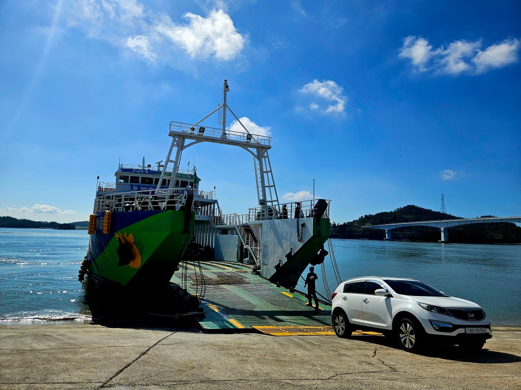 Un ferry coloré à quai dans un terminal presque vide, avec une voiture blanche stationnée à proximité et un ciel bleu en arrière-plan.