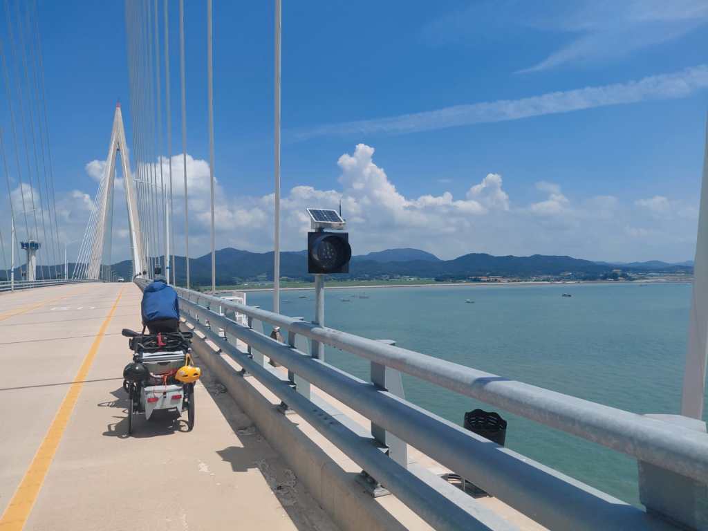 Une vue d'un cycliste sur un pont moderne, avec des câbles de suspension et des montagnes en arrière-plan sous un ciel bleu clair.