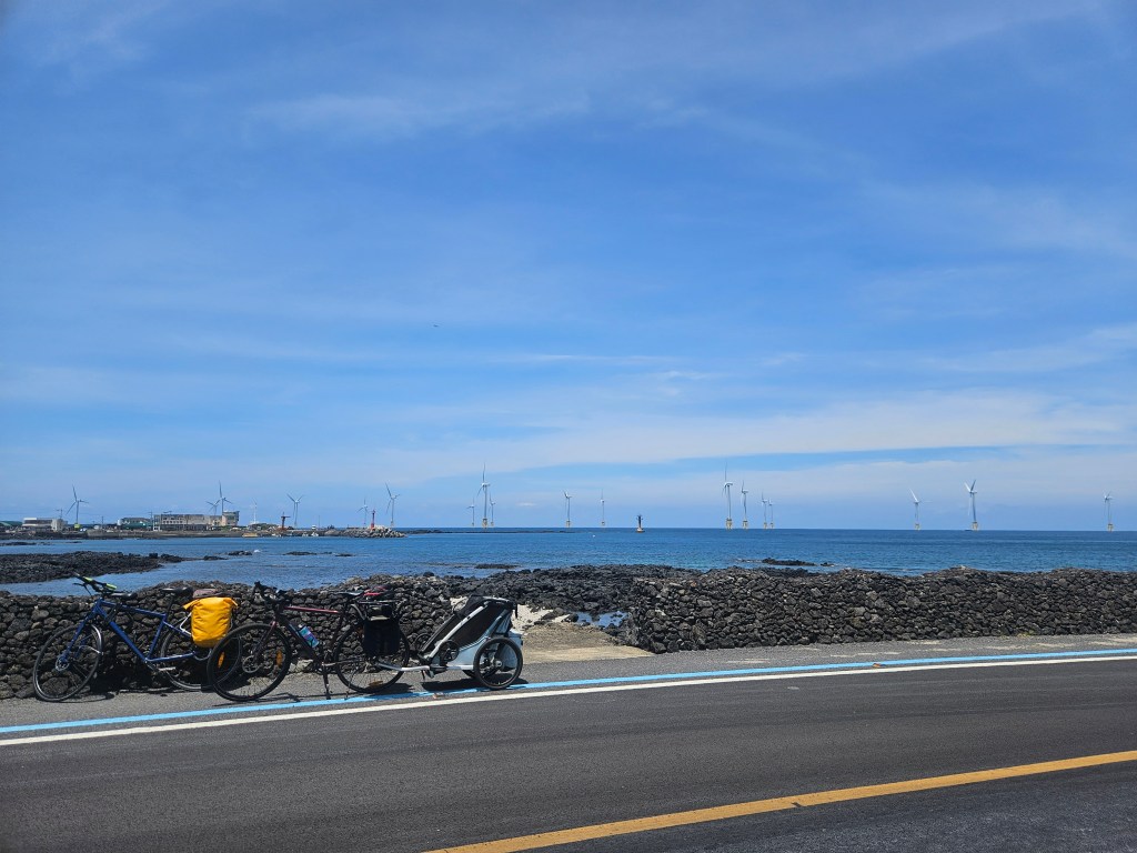 Un paysage côtier de Jeju, Corée du Sud, avec des vélos garés sur le bord de la route. Des éoliennes sont visibles au loin sur la mer. Le ciel est dégagé et bleu.