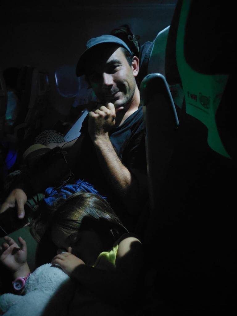 Un homme souriant, assis dans un avion, tenant un écouteur, avec une jeune fille endormie sur son épaule.