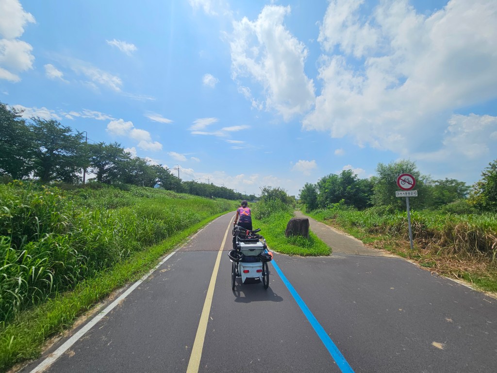 Une scène d'une véloroute en Corée du Sud avec deux cyclistes devant, une remorque de vélo, des espaces verts de chaque côté, et un ciel bleu parsemé de nuages.