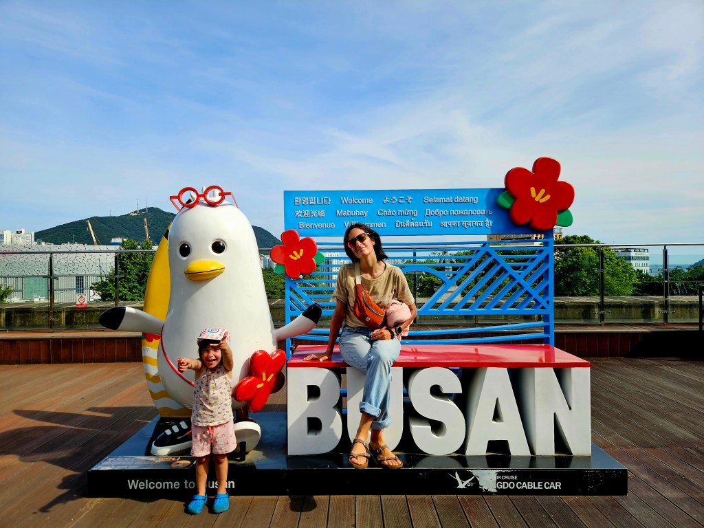 Une petite fille souriante posant avec une mascotte en forme de pingouin, devant un panneau 'Welcome to Busan', avec des fleurs colorées en arrière-plan.