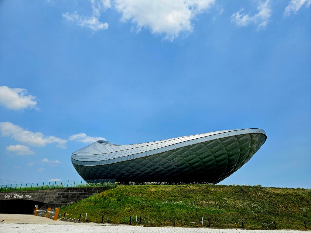 Vue d'un bâtiment moderne en forme de bateau, situé sur une colline verdoyante, sous un ciel bleu avec quelques nuages.