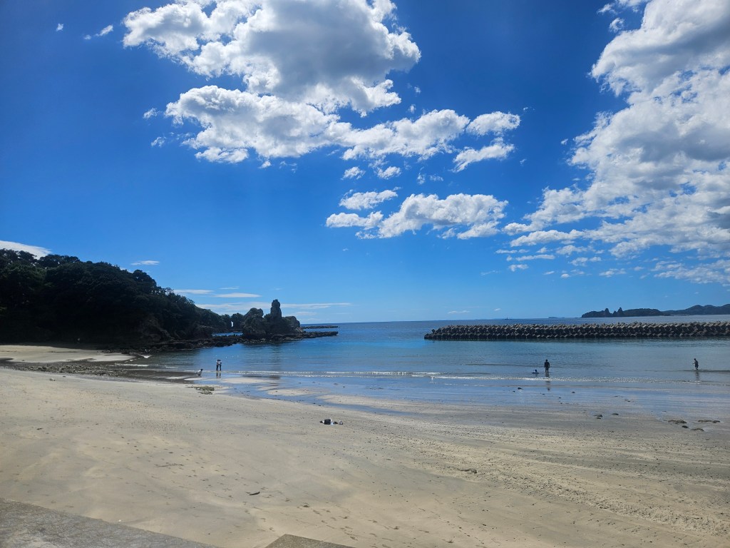 Plage calme avec des eaux cristallines, entourée de petites falaises et de verdure, sous un ciel bleu parsemé de nuages.