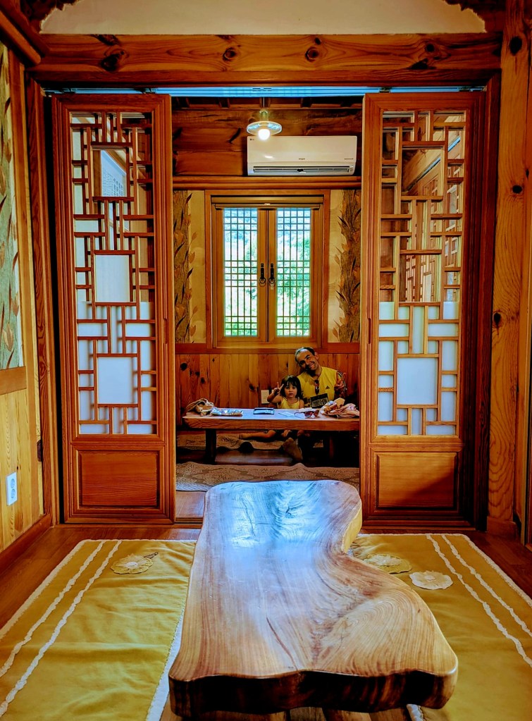 Intérieur d'une maison traditionnelle coréenne (hanok) avec un homme et un enfant assis à une petite table basse, entourés de murs en bois et de fenêtres à treillis.