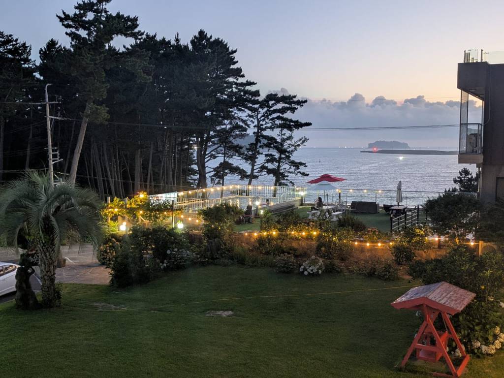 Vue d'un espace extérieur au bord de la mer, avec des arbres au fond, des lumières scintillantes, un mobilier de jardin, et un petit bâtiment contemporain.