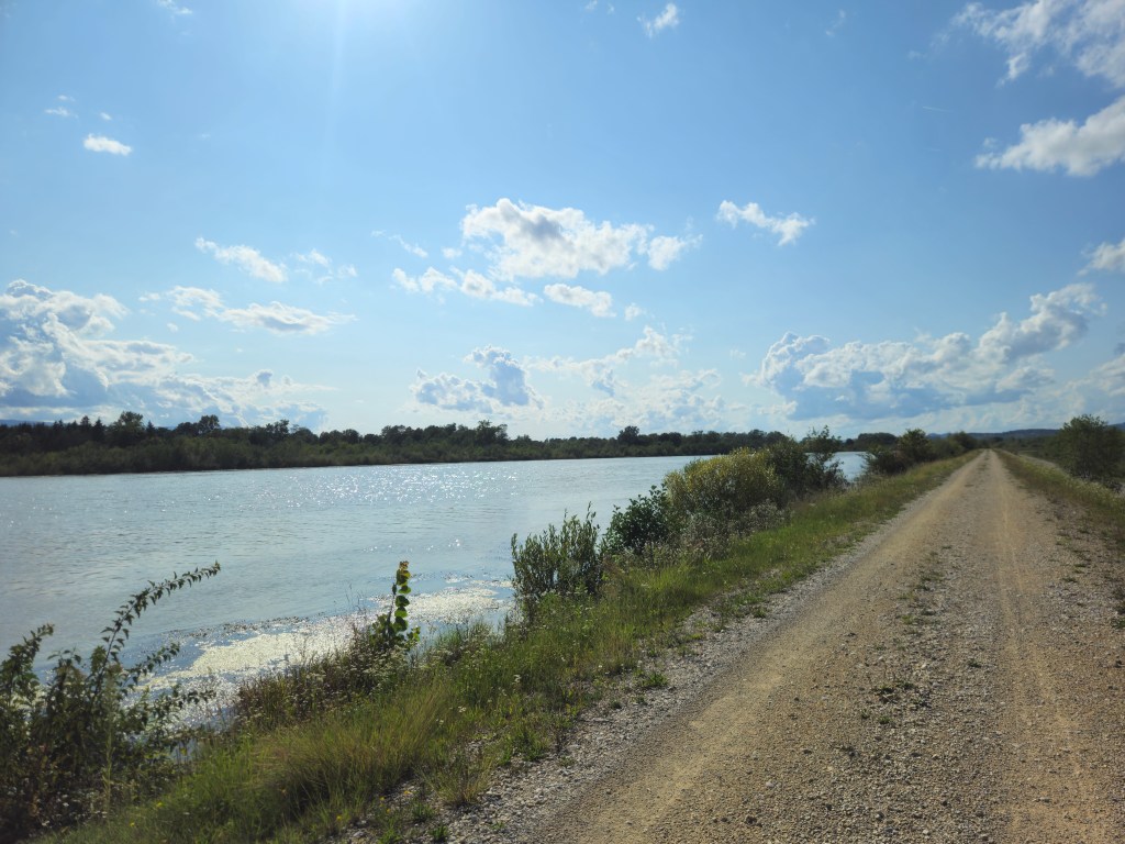 Un chemin de terre longe un cours d'eau, avec des herbes et des buissons sur les bords, sous un ciel partiellement nuageux.
