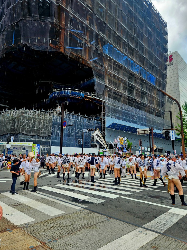 Des hommes en tenue traditionnelle défilent à Fukuoka, lors d'un festival, tandis qu'un bâtiment en rénovation est visible en arrière-plan.