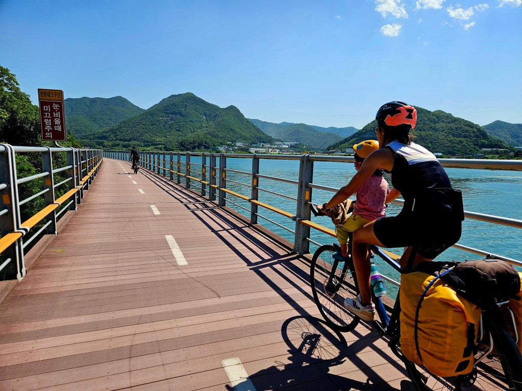Une mère et son enfant circulent à vélo sur une piste cyclable au bord d'un lac, entourés de collines verdoyantes et sous un ciel bleu ensoleillé.