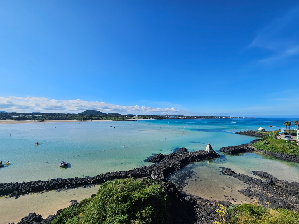 Vue panoramique de la plage de Sogeummak à Jeju, avec une mer claire et peu profonde, des formations rocheuses noires au premier plan et des personnes se baignant au loin sous un ciel bleu.