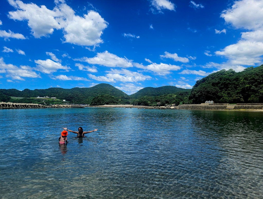 Deux personnes se baignent dans une eau claire, entourées de collines verdoyantes et d'un ciel bleu parsemé de nuages.