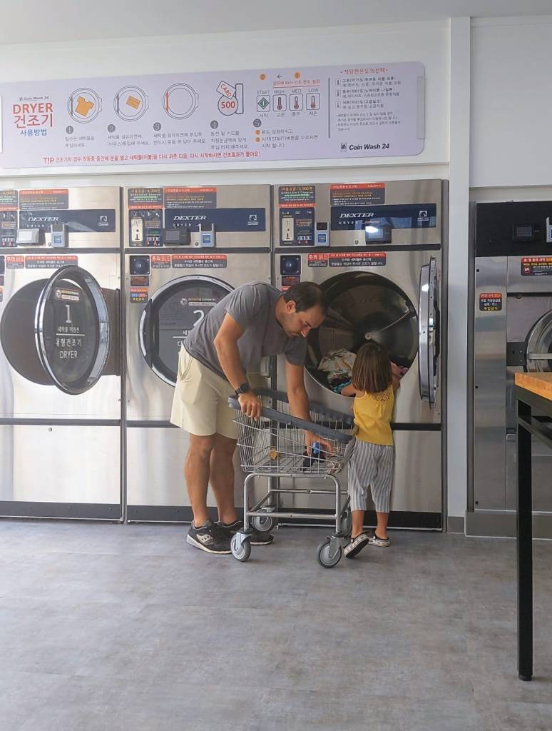 Un adulte et un enfant s'apprêtent à utiliser un sèche-linge dans une laverie automatique. Le sèche-linge est en métal avec une porte transparente, et l'adulte aide l'enfant à placer des vêtements à l'intérieur. On aperçoit aussi un chariot à linge à côté d'eux.