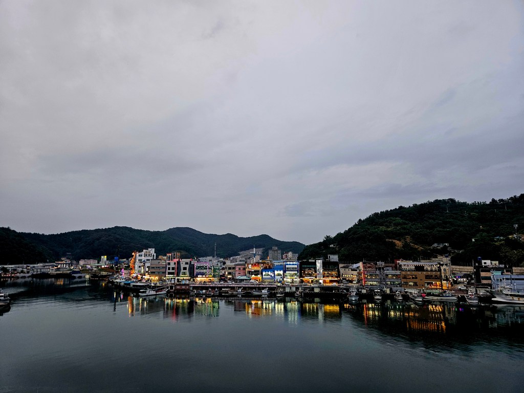 Vue panoramique d'une ville portuaire coréenne avec des bâtiments illuminés le long de la côte, reflétés dans l'eau calme sous un ciel nuageux.