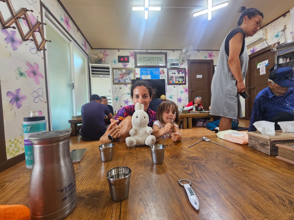 Une mère et sa fille dans un restaurant coréen, avec une peluche au centre, entourées de clients assis à des tables basses.