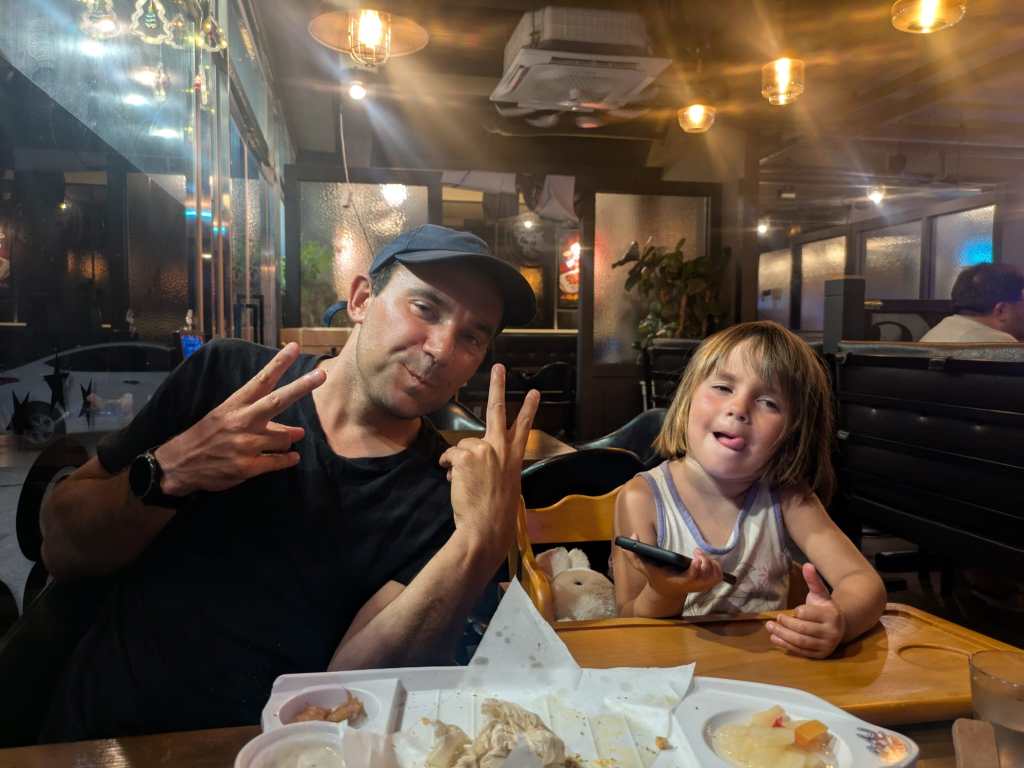 Un homme et un enfant à une table de restaurant, posant pour la photo avec des gestes de paix et des sourires, avec des plats sur la table en arrière-plan.