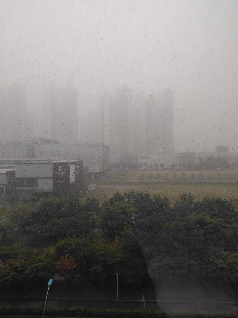 Vue nébuleuse d'un paysage urbain sous une pluie intense, avec des immeubles en arrière-plan flous, vus à travers une fenêtre mouillée.