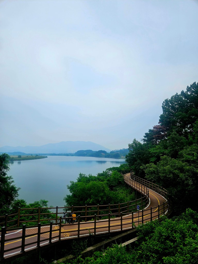 Vue panoramique d'une véloroute serpentant le long d'un plan d'eau, entourée de verdure et de collines, avec un ciel nuageux.