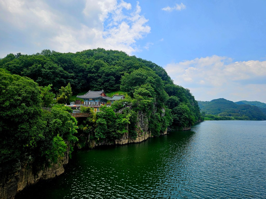 Vue d'une maison traditionnelle coréenne perchée sur une falaise verdoyante surplombant un lac calme sous un ciel nuageux.