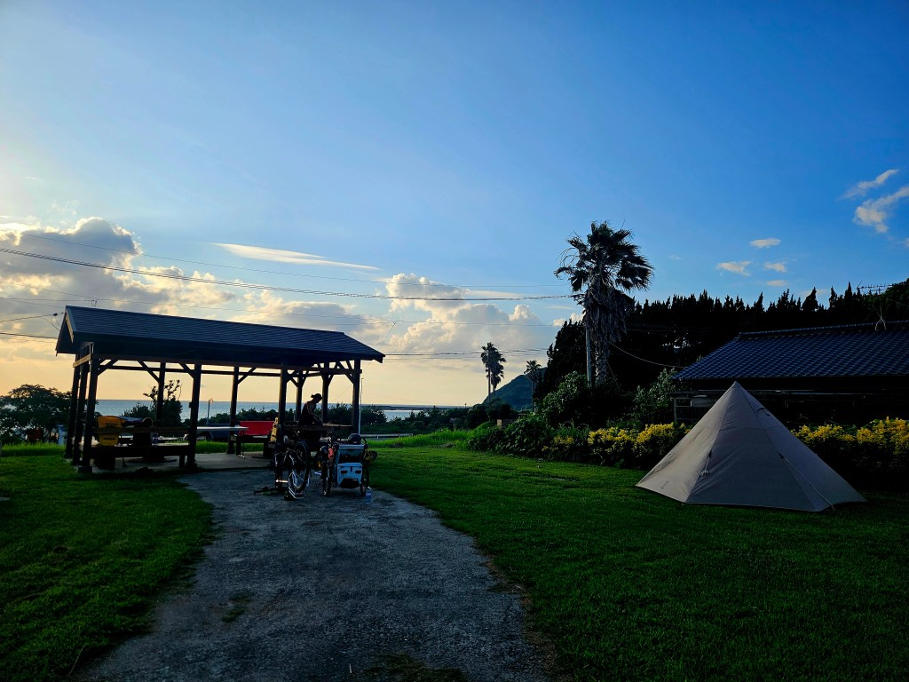 Un camping en pleine nature avec une tente, des vélos garés et un abri en bois sous un ciel bleu.