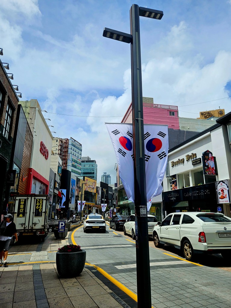 Rue animée en Corée du Sud, avec des vêtements colorés et des drapeaux sud-coréens accrochés aux lampadaires, entourée de magasins et de véhicules.