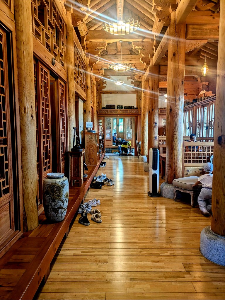 Intérieur d'un hanok traditionnel coréen, montrant un couloir en bois avec des chaussures laissées sur le sol, des fenêtres à panneaux de bois et un éclairage naturel.