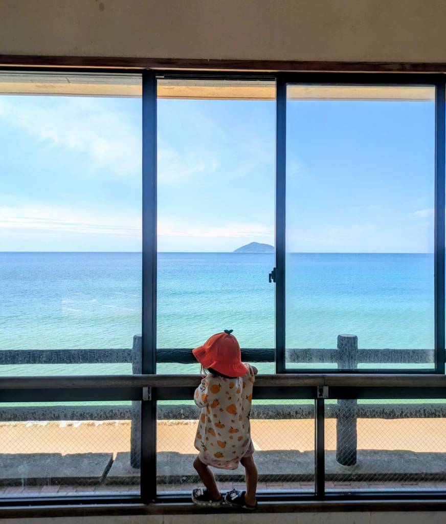 Un enfant avec un chapeau orange se tient debout devant une grande fenêtre, regardant la mer paisible et une petite île au loin.