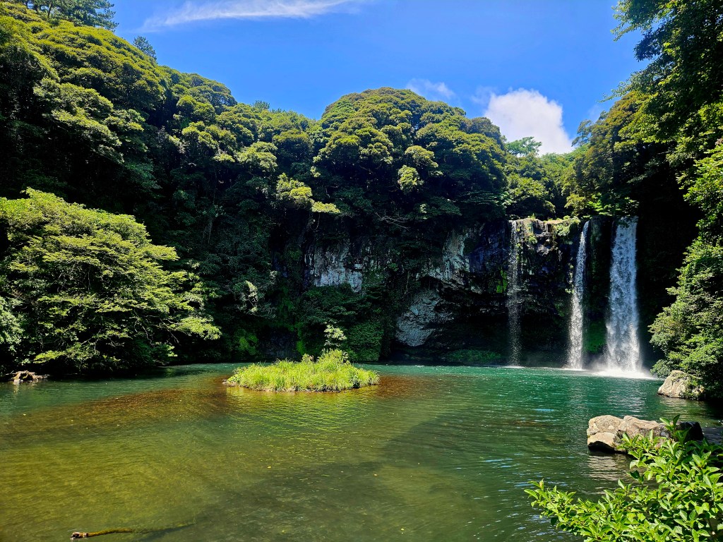Vue de la cascade de Cheonjiyeon, entourée de verdure, tombant dans un bassin d'eau claire.