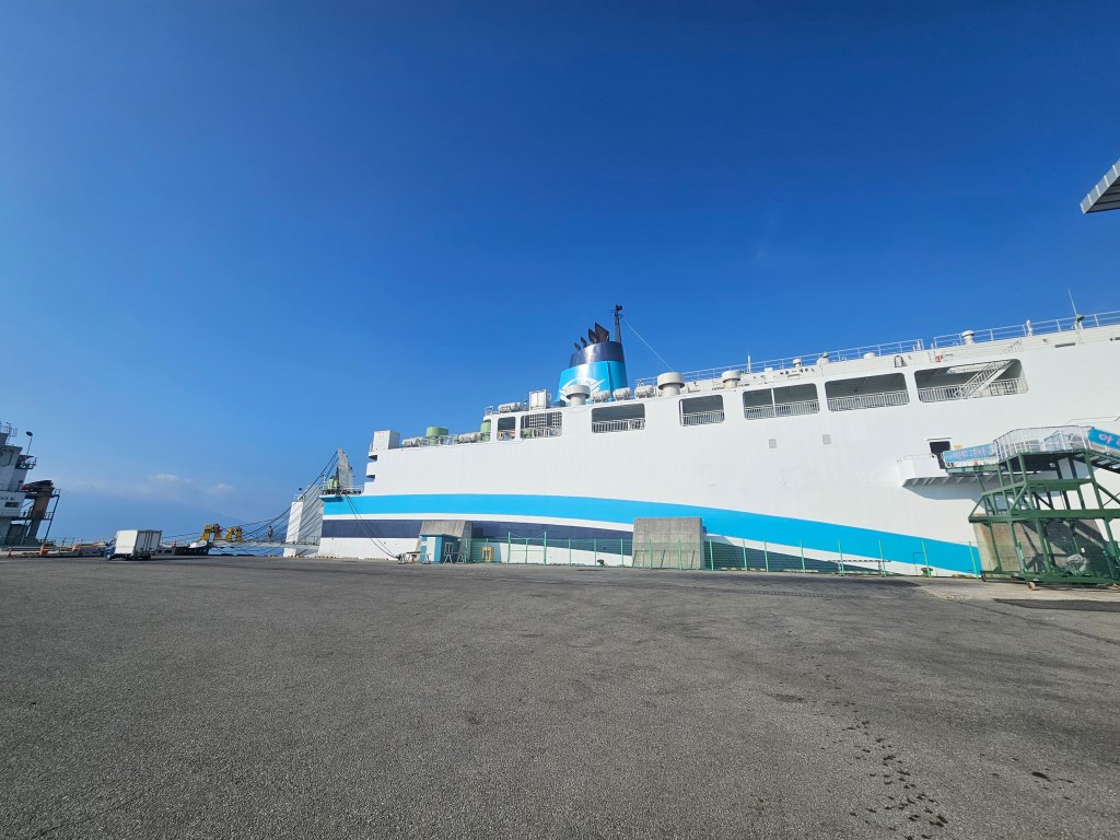 Un immense ferry amarré au port sous un ciel bleu, avec une rampe d'accès visible et un quai en béton.